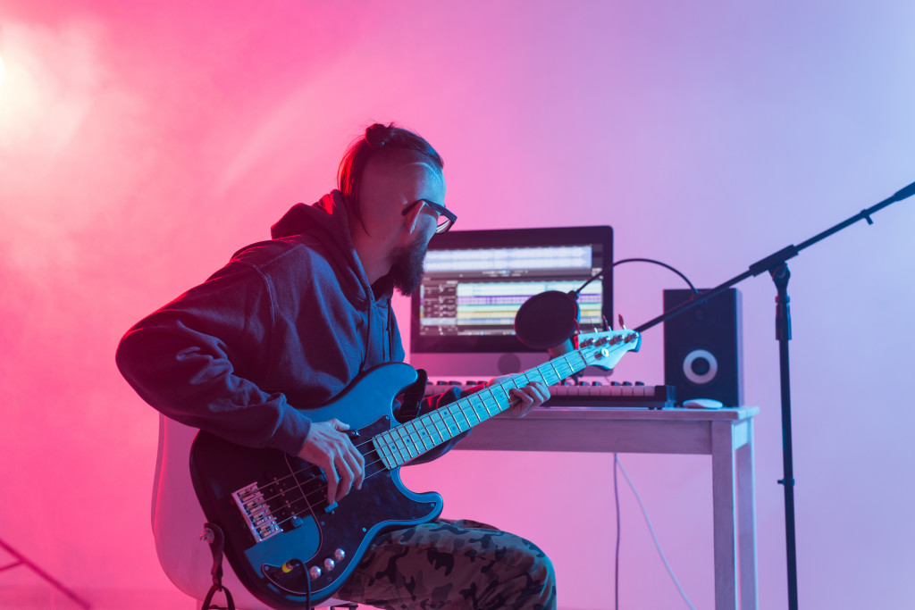 A man plays a bass guitar in front of a mic, condenser, and computer at their home studio